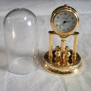 May include: A gold-colored quartz clock with a white face and black numerals, labeled "GENEVA QUARTZ." The clock sits on a decorative base with a clear glass dome beside it. The clock's design includes a pendulum-like feature.
