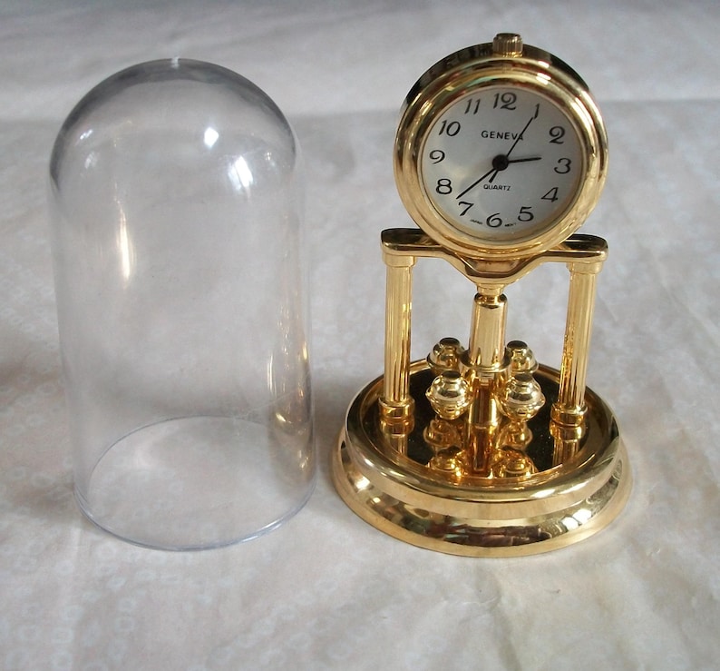 May include: A gold-colored quartz clock with a white face and black numerals, labeled "GENEVA QUARTZ." The clock sits on a decorative base with a clear glass dome beside it. The clock's design includes a pendulum-like feature.