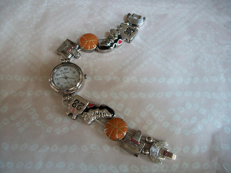 May include: A silver watch with a white face and black hands. The watch band is decorated with basketballs, sneakers, and the number 23.