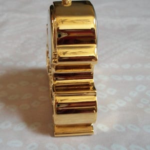May include: A gold-colored, rectangular-shaped watch with a ridged band and a small, circular crown. The watch has a shiny, reflective surface and a white watch face. The watch is a classic accessory.