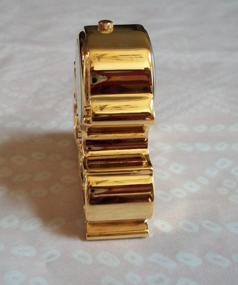 May include: A gold-colored, rectangular-shaped watch with a ridged band and a small, circular crown. The watch has a shiny, reflective surface and a white watch face. The watch is a classic accessory.