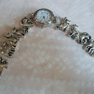 May include: A silver watch with a white face and black numbers. The watch band is made of a series of linked silver cat figurines.