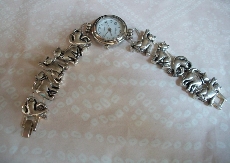 May include: A silver watch with a white face and black numbers. The watch band is made of a series of linked silver cat figurines.