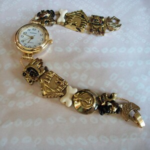 May include: Gold-tone watch with a white face and black numbers. The watch band is made of a chain with dog-themed charms, including a dog house, bones, and dogs.