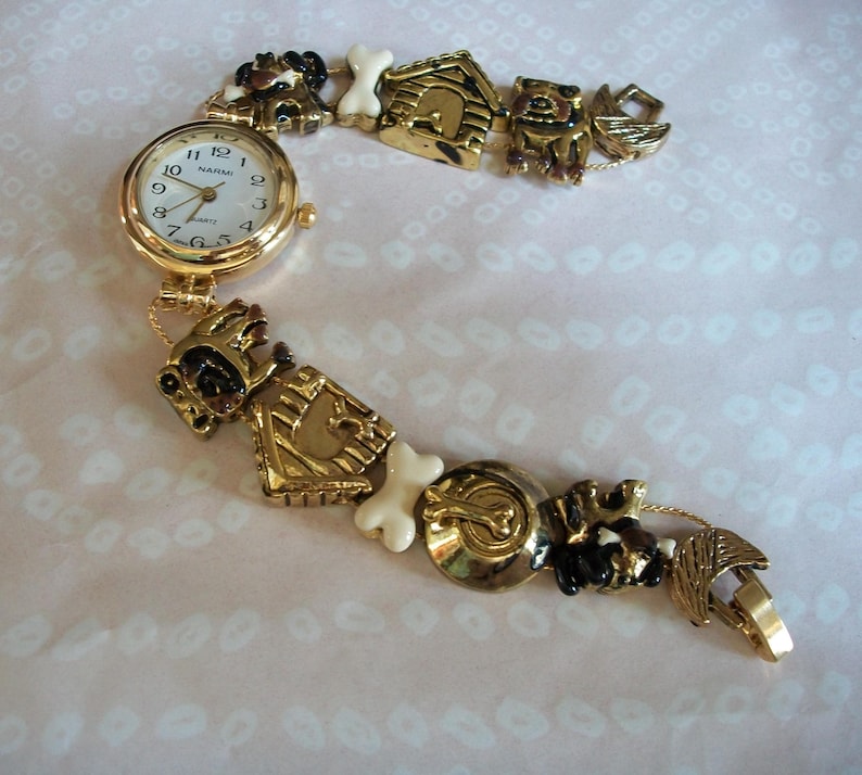 May include: Gold-tone watch with a white face and black numbers. The watch band is made of a chain with dog-themed charms, including a dog house, bones, and dogs.