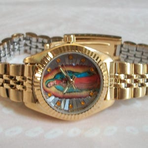 May include: A gold-tone wristwatch with a gold-tone metal band. The watch face features a colorful image of the Virgin Mary.