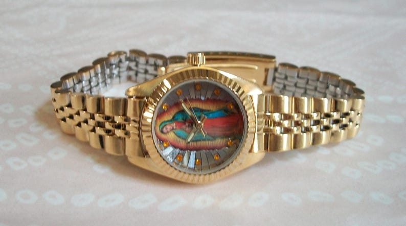 May include: A gold-tone wristwatch with a gold-tone metal band. The watch face features a colorful image of the Virgin Mary.