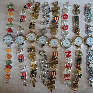 May include: A collection of ten charm bracelet watches with different colored charms. The charms include flags, animals, fruits, and letters. The watches have silver bands and white faces.
