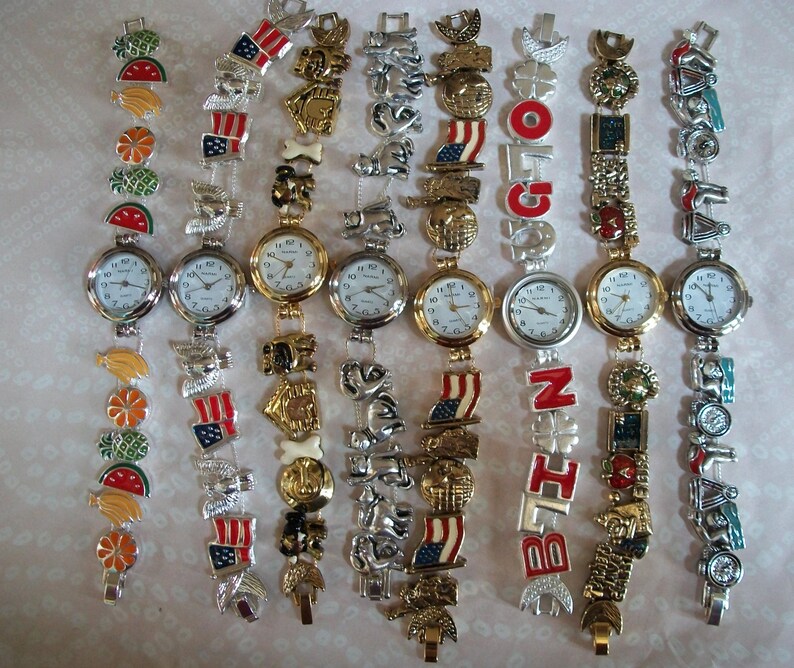 May include: A collection of ten charm bracelet watches with different colored charms. The charms include flags, animals, fruits, and letters. The watches have silver bands and white faces.