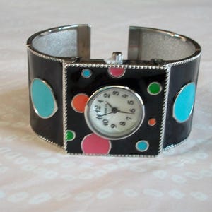 May include: A black cuff bracelet watch with a white face and colorful polka dots. The watch face has black Roman numeral hour markers and black hands.