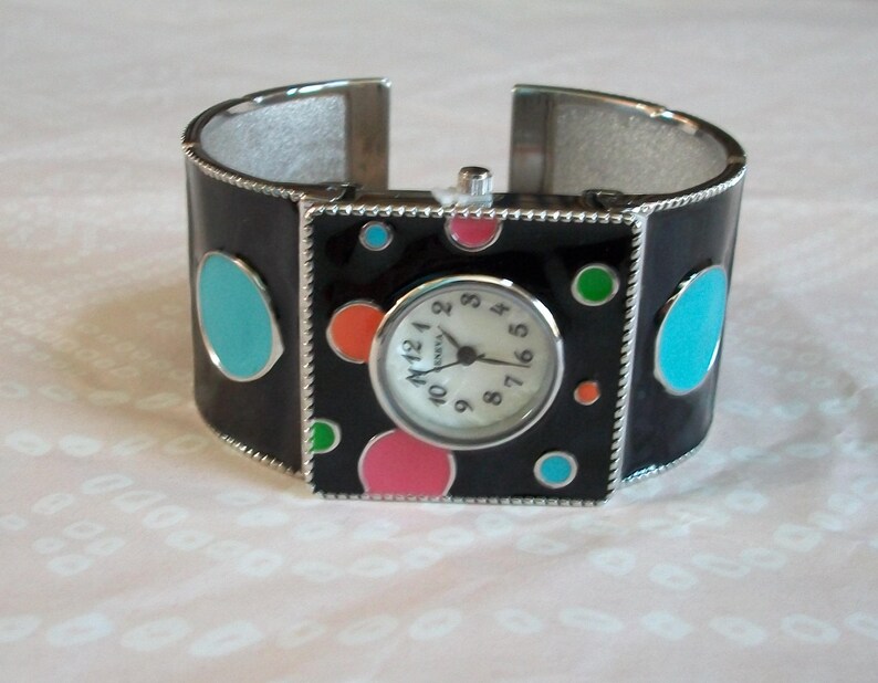 May include: A black cuff bracelet watch with a white face and colorful polka dots. The watch face has black Roman numeral hour markers and black hands.