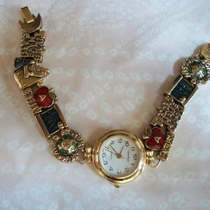 May include: Gold-tone wristwatch with a white face and black numbers. The watch band is a charm bracelet with various school-themed charms, including apples, books, and a globe.