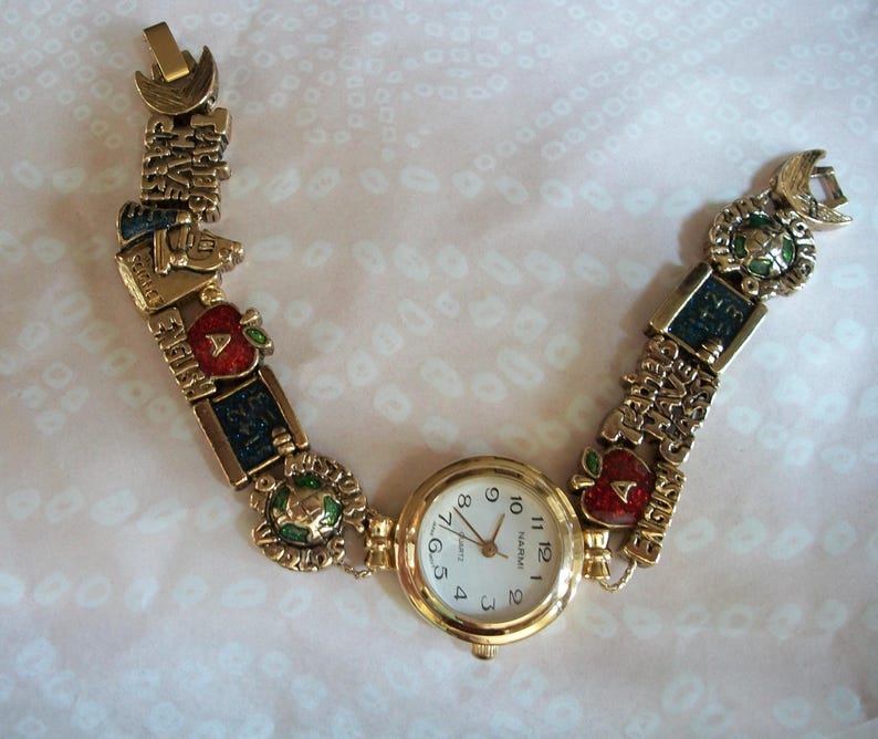 May include: Gold-tone wristwatch with a white face and black numbers. The watch band is a charm bracelet with various school-themed charms, including apples, books, and a globe.