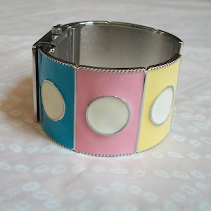 May include: A silver bangle bracelet with three colored panels. The panels are turquoise, pink, and yellow, each with a white circle in the center.