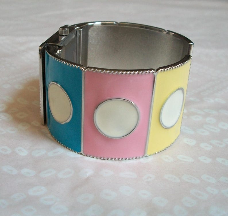 May include: A silver bangle bracelet with three colored panels. The panels are turquoise, pink, and yellow, each with a white circle in the center.
