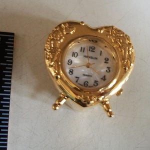 May include: A gold-colored, heart-shaped quartz clock with a white face and black numerals. The clock has a decorative floral design around the face and stands on three small legs. The clock face reads "Hanslin Quartz".