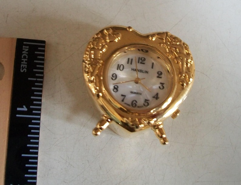 May include: A gold-colored, heart-shaped quartz clock with a white face and black numerals. The clock has a decorative floral design around the face and stands on three small legs. The clock face reads "Hanslin Quartz".