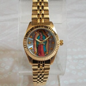 May include: A gold-tone wristwatch with a round face featuring a colorful image of the Virgin Mary. The watch has a gold-tone metal band.