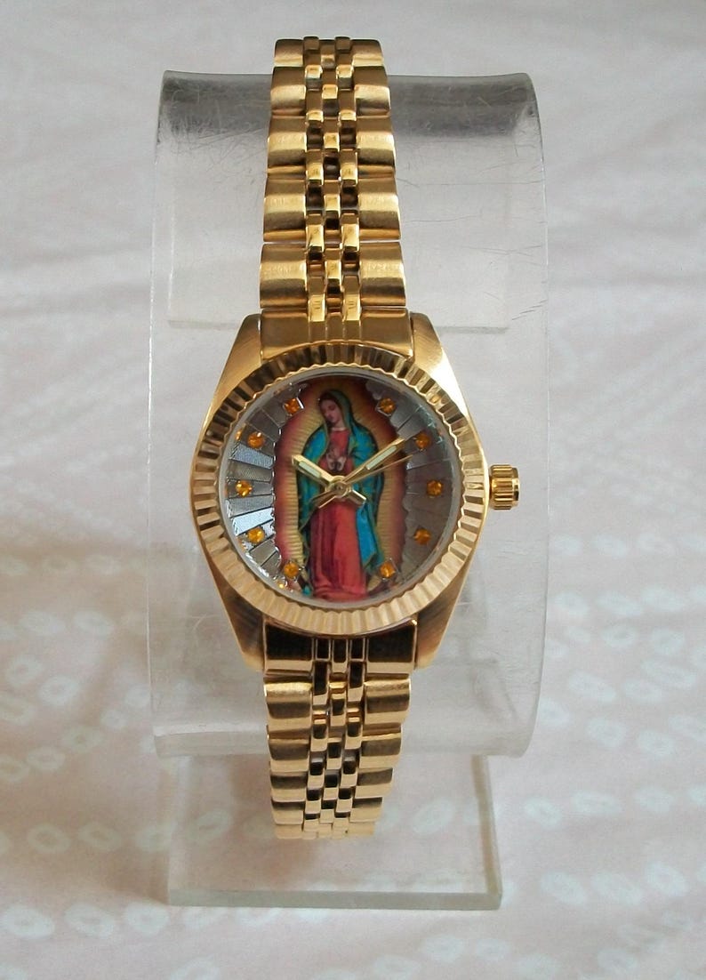 May include: A gold-tone wristwatch with a round face featuring a colorful image of the Virgin Mary. The watch has a gold-tone metal band.