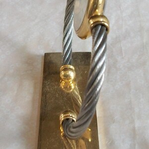 May include: A gold-toned watch with a silver-colored face and a twisted silver and gold band. The watch is mounted on a rectangular gold-colored base. The watch has a classic, elegant design.