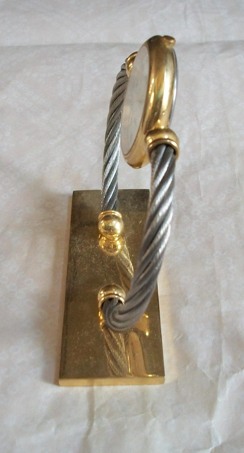 May include: A gold-toned watch with a silver-colored face and a twisted silver and gold band. The watch is mounted on a rectangular gold-colored base. The watch has a classic, elegant design.