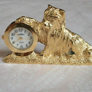 May include: A gold-colored dog-shaped clock with a round clock face. The clock face has black numerals and hands, and the word "Quartz" is printed on the face. The clock is a decorative item.