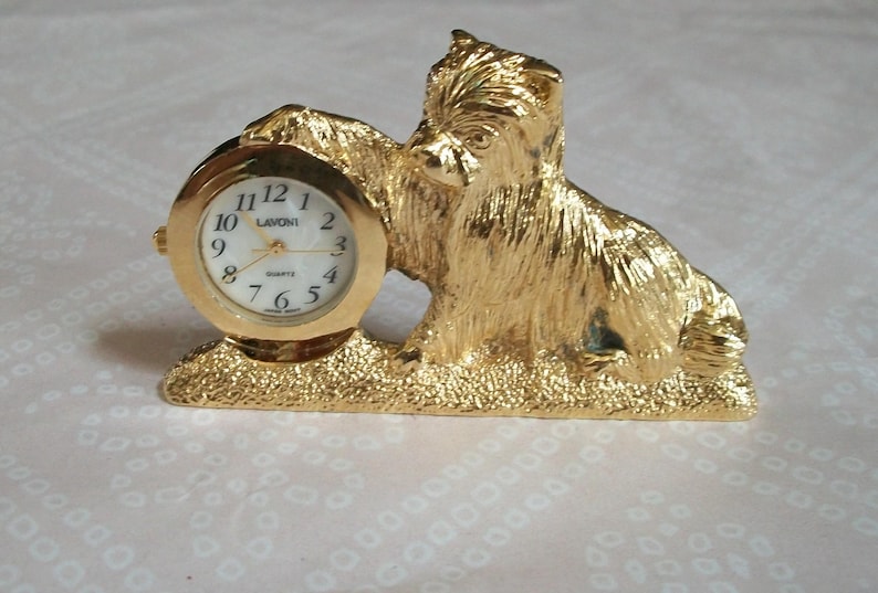 May include: A gold-colored dog-shaped clock with a round clock face. The clock face has black numerals and hands, and the word "Quartz" is printed on the face. The clock is a decorative item.