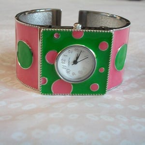 May include: A pink and green polka dot cuff bracelet watch with a white face and silver hands. The watch face says "Geneva" and has black numbers.