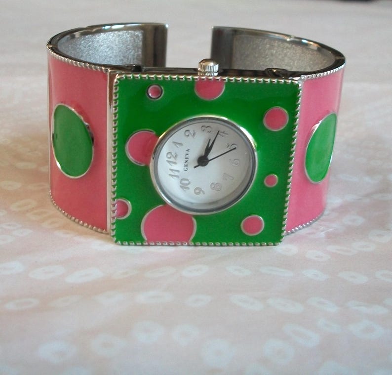 May include: A pink and green polka dot cuff bracelet watch with a white face and silver hands. The watch face says "Geneva" and has black numbers.