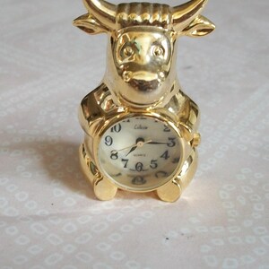 May include: A gold-colored bull-shaped clock with a round clock face. The clock face has black numerals and hands. The clock is a collectible item.