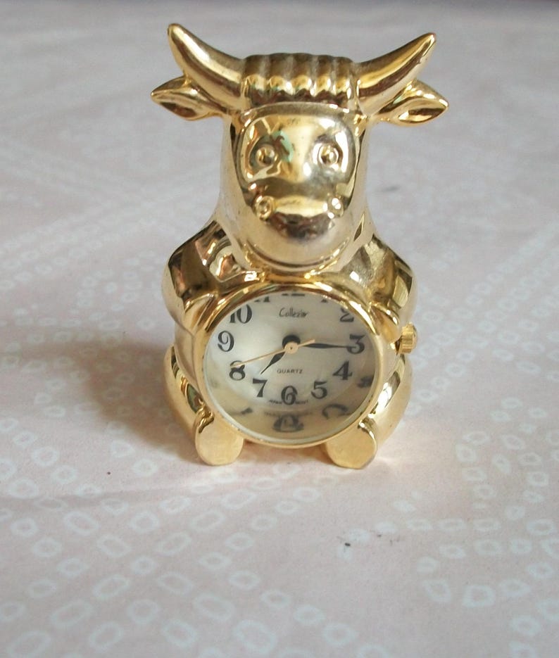 May include: A gold-colored bull-shaped clock with a round clock face. The clock face has black numerals and hands. The clock is a collectible item.