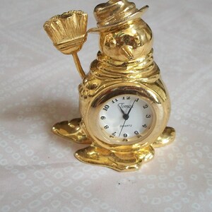 May include: A gold-colored snowman-shaped quartz clock with a white clock face. The clock has a hat and holds a broom. The clock face reads "Timex Quartz".