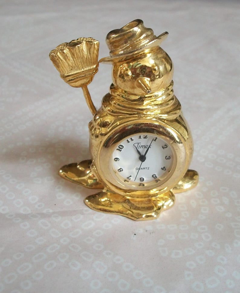 May include: A gold-colored snowman-shaped quartz clock with a white clock face. The clock has a hat and holds a broom. The clock face reads "Timex Quartz".