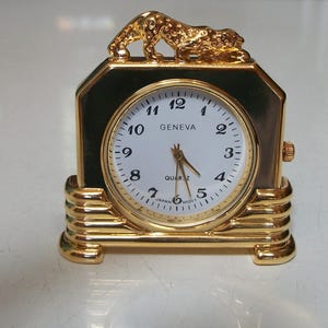 May include: A gold-colored Geneva quartz clock with a white face and black numerals. The clock has a rectangular shape with a decorative gold leopard on top. The clock is approximately 4 inches tall.