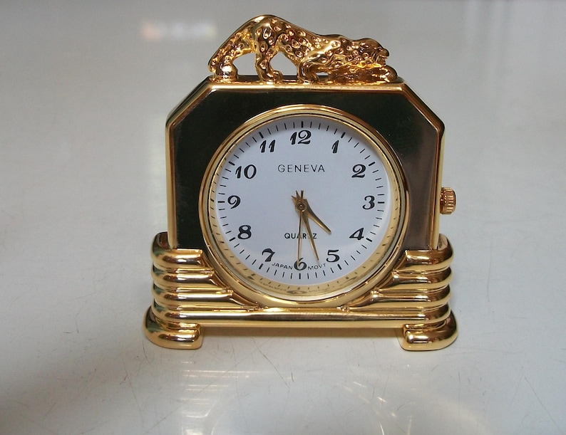 May include: A gold-colored Geneva quartz clock with a white face and black numerals. The clock has a rectangular shape with a decorative gold leopard on top. The clock is approximately 4 inches tall.