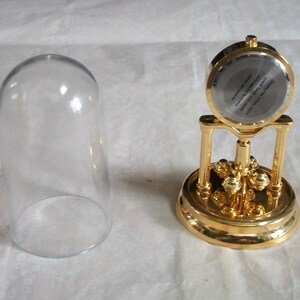 May include: A gold-colored, decorative clock with a clear glass dome. The clock has a round face and a detailed base. The glass dome is separate from the clock. The clock's face has text that is difficult to read.