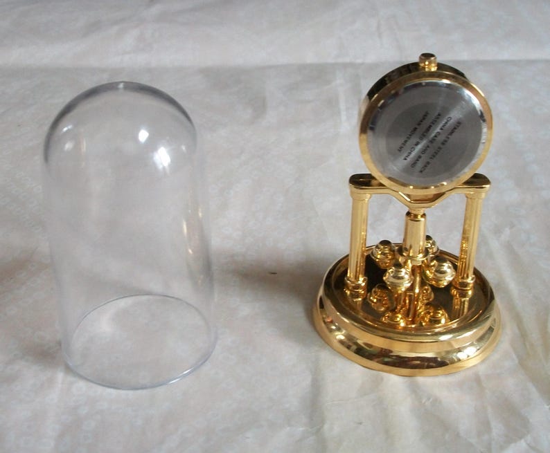 May include: A gold-colored, decorative clock with a clear glass dome. The clock has a round face and a detailed base. The glass dome is separate from the clock. The clock's face has text that is difficult to read.