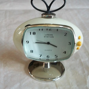 May include: A retro-style, off-white quartz clock with a silver base and accents. The clock face is rectangular with black numerals and the words "URBAN STATION" printed on it. A black loop design sits atop the clock.