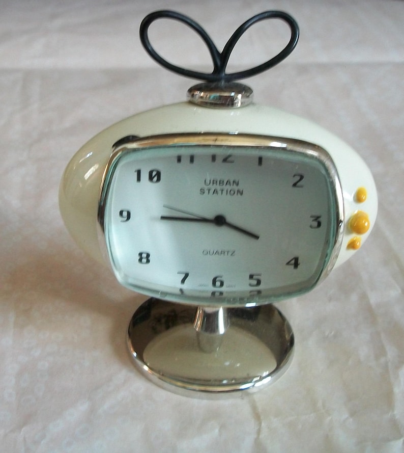 May include: A retro-style, off-white quartz clock with a silver base and accents. The clock face is rectangular with black numerals and the words "URBAN STATION" printed on it. A black loop design sits atop the clock.