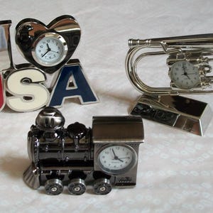 May include: A set of four decorative clocks featuring a silver train engine, a silver trumpet, a silver heart with the word "USA" and a silver heart with the words "I Love" on it.