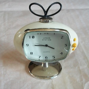 May include: A retro-style quartz clock with a cream-colored, oval-shaped body and a silver base. The clock face is rectangular with black numerals and hands, and the text "URBAN STATION" is visible. A black, looped handle sits atop the clock.
