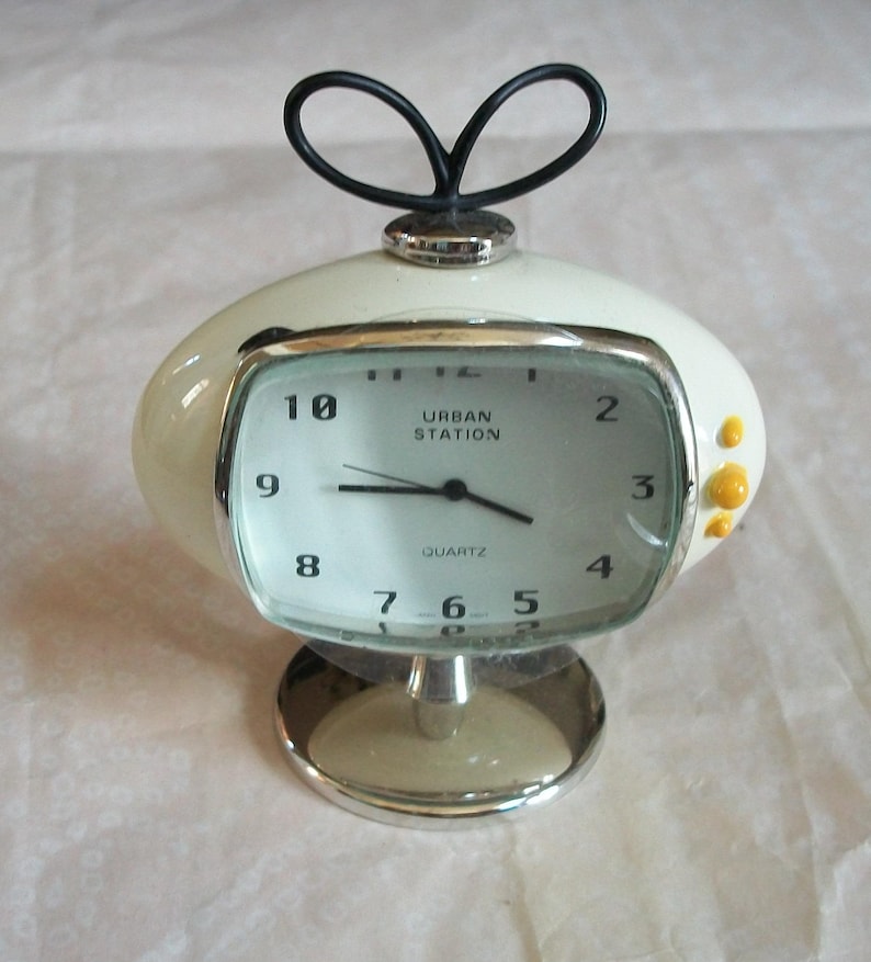 May include: A retro-style quartz clock with a cream-colored, oval-shaped body and a silver base. The clock face is rectangular with black numerals and hands, and the text "URBAN STATION" is visible. A black, looped handle sits atop the clock.