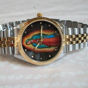 May include: A silver and gold tone wristwatch with a black face featuring a colorful image of the Virgin Mary. The watch has a stainless steel band with gold accents.
