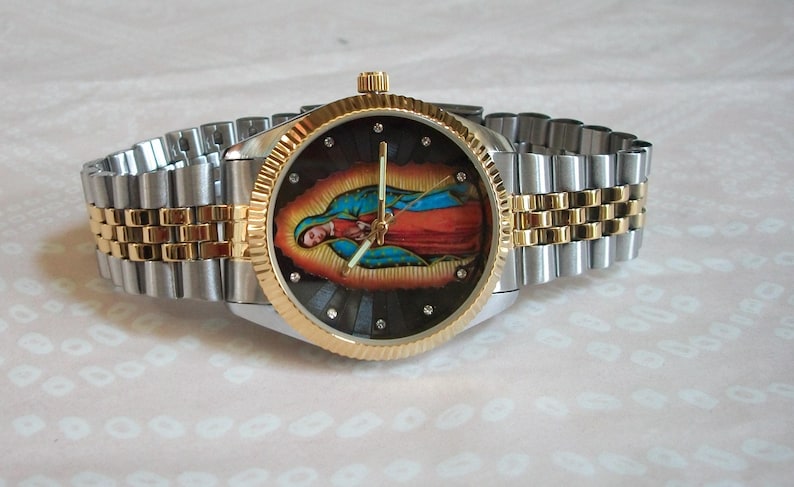 May include: A silver and gold tone wristwatch with a black face featuring a colorful image of the Virgin Mary. The watch has a stainless steel band with gold accents.