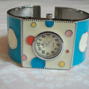 May include: A silver cuff bracelet watch with a white face and colorful polka dots. The watch face says "Demacci Quartz".