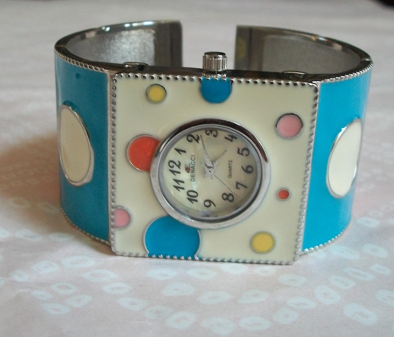 May include: A silver cuff bracelet watch with a white face and colorful polka dots. The watch face says "Demacci Quartz".