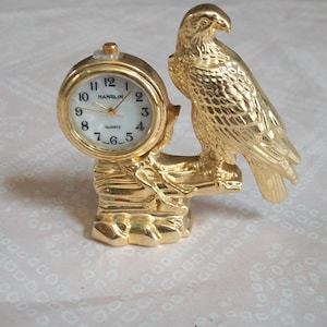 May include: A gold-colored quartz clock with a white face and black numerals, featuring the brand name "HANSLIN." The clock is integrated into a decorative gold eagle figurine perched on a rock formation. The clock's face is round.