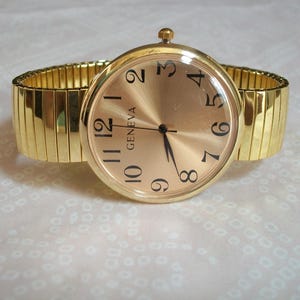 May include: A gold-tone wristwatch featuring a round face with a metallic gold dial, black Arabic numerals, and the brand name Geneva printed in black. The watch includes a flexible, expandable gold-tone metal link band with a polished finish.