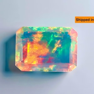 May include: A multi-coloured rectangular gemstone with a faceted cut, displaying hues of green, orange, and blue. The gem is set against a pale blue backdrop. The text "Shipped in 24 hrs" is visible in the top right corner.
