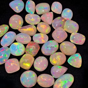 Può includere: Una collezione di opali iridescenti di varie forme, tra cui ovali, rotondi e a goccia. Gli opali mostrano una gamma di colori, tra cui rosa, giallo, blu e verde, con un effetto cangiante e multicolore. Gli opali sono disposti su uno sfondo nero.